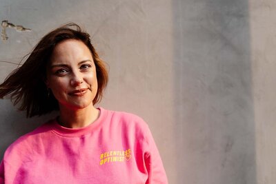 Bio photo of Charlie Flounders holding a yellow bag and her camera, standing in front of a yellow door. She holds her camera to depict that she is a photographer